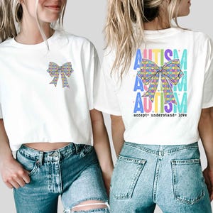 May include: White t-shirts with a colorful bow design. One shirt has the bow on the front, the other on the back with the word "AUTISM" in multiple colors and the words "accept- understand- love".