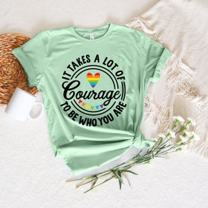May include: A light green t-shirt with the text "IT TAKES A LOT OF Courage TO BE WHO YOU ARE" in black. The word "Courage" is in a circle with a rainbow heart and rainbow triangles. A white mug and flowers are in the background.