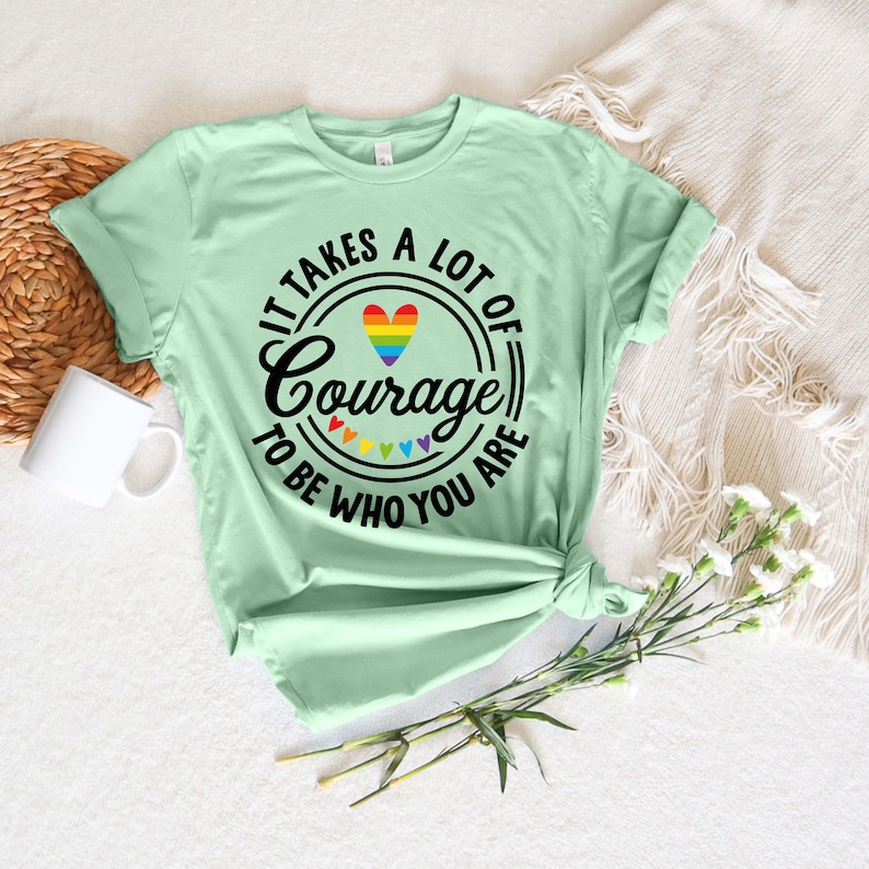 May include: A light green t-shirt with the text "IT TAKES A LOT OF Courage TO BE WHO YOU ARE" in black. The word "Courage" is in a circle with a rainbow heart and rainbow triangles. A white mug and flowers are in the background.