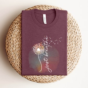May include: A maroon t-shirt featuring a dandelion graphic and the words "just breathe" in white script. The dandelion design has a colourful watercolour effect. The shirt is folded on a woven, circular placemat.