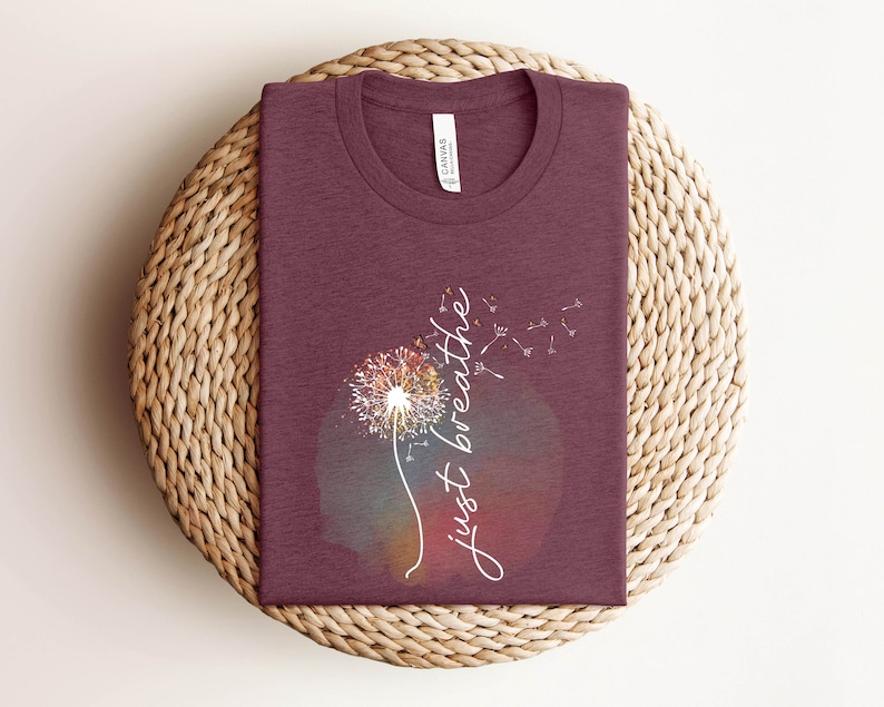 May include: A maroon t-shirt featuring a dandelion graphic and the words "just breathe" in white script. The dandelion design has a colourful watercolour effect. The shirt is folded on a woven, circular placemat.