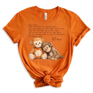 K&ouml;nnte beinhalten: Orangefarbenes T-Shirt mit einer Zeichnung von zwei Pl&uuml;schaffen. Einer ist braun und beige, der andere ist braun. Das Shirt hat handgeschriebenen Text, der lautet: "Dear Peach, I am not breath, art a beating heart...Love, Your Plush."