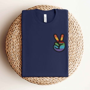 K&ouml;nnte beinhalten: Ein marineblaues T-Shirt mit einem gestickten Regenbogen-Friedenszeichen auf der linken Brust.