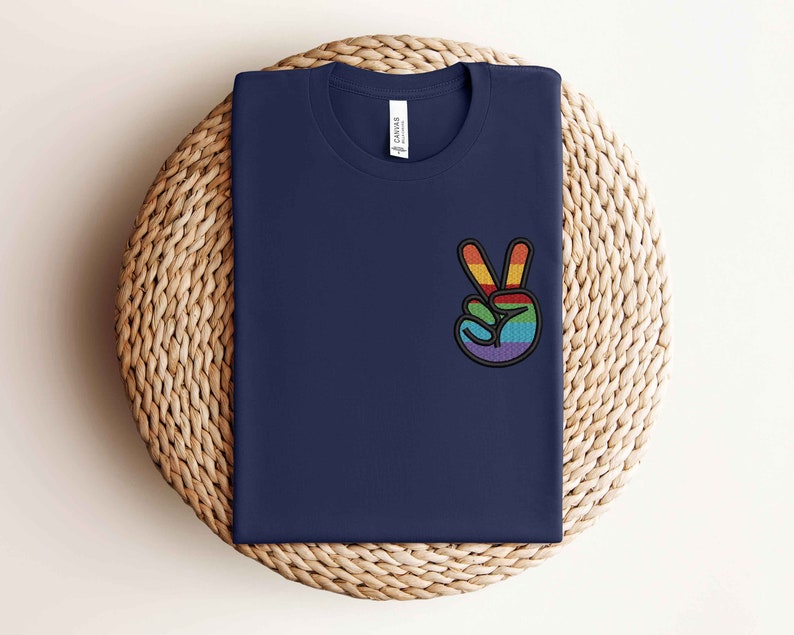 K&ouml;nnte beinhalten: Ein marineblaues T-Shirt mit einem gestickten Regenbogen-Friedenszeichen auf der linken Brust.