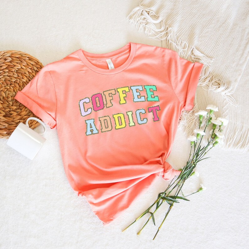 May include: A peach colored t-shirt with the words "COFFEE ADDICT" in colorful letters. The letters are outlined in gold glitter.