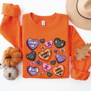 Puede incluir: Sudadera naranja con un dise&ntilde;o de tem&aacute;tica de Halloween con corazones de caramelo con frases espeluznantes como "Haunt Me", "Hey Boo", "Spooky Vibes" y "Ghoul Crush".