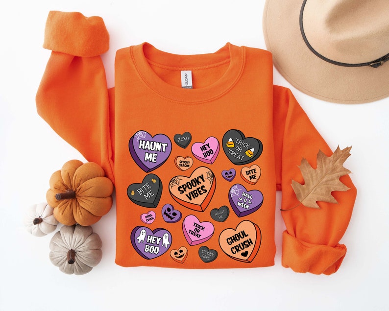 Puede incluir: Sudadera naranja con un dise&ntilde;o de tem&aacute;tica de Halloween con corazones de caramelo con frases espeluznantes como "Haunt Me", "Hey Boo", "Spooky Vibes" y "Ghoul Crush".