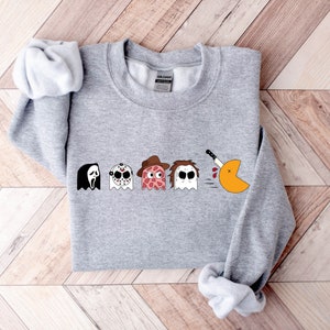 Pode incluir: Camisola de cor cinzenta com um gr&aacute;fico de Pac-Man. O Pac-Man est&aacute; a ser perseguido por quatro fantasmas, cada um com uma m&aacute;scara de um personagem de filme de terror diferente: Ghostface, Jason Voorhees, Freddy Krueger e Leatherface.
