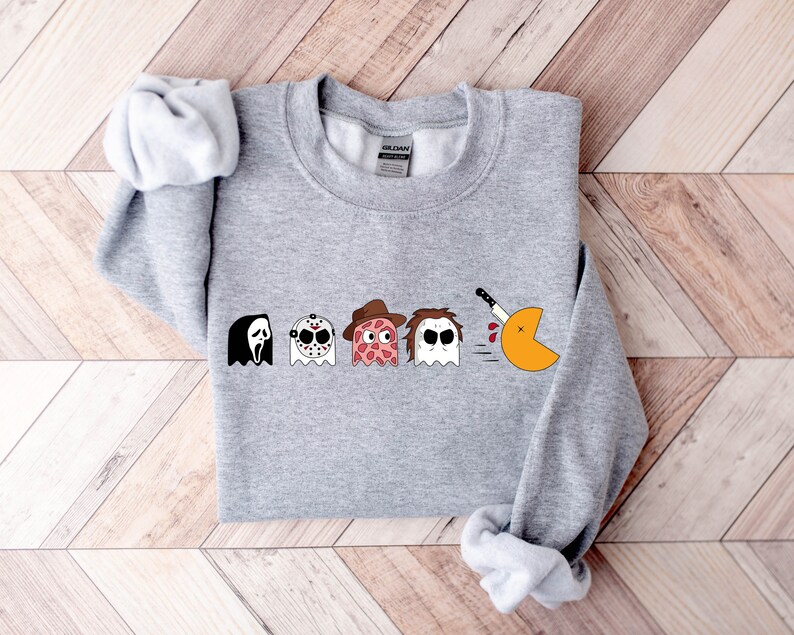 Pode incluir: Camisola de cor cinzenta com um gr&aacute;fico de Pac-Man. O Pac-Man est&aacute; a ser perseguido por quatro fantasmas, cada um com uma m&aacute;scara de um personagem de filme de terror diferente: Ghostface, Jason Voorhees, Freddy Krueger e Leatherface.
