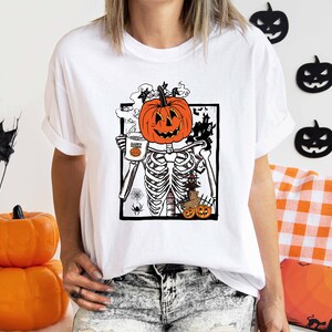 K&ouml;nnte beinhalten: Wei&szlig;es T-Shirt mit einem Grafikdruck eines Skeletts, das eine Tasse mit dem Text "Happy" und einen K&uuml;rbis-Kopf h&auml;lt. Das Skelett ist von Flederm&auml;usen und einem Spukhaus umgeben.