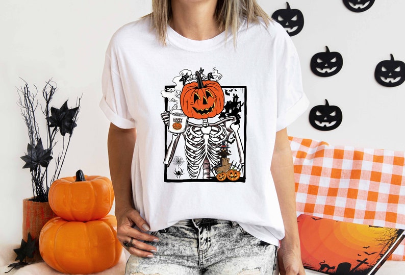 K&ouml;nnte beinhalten: Wei&szlig;es T-Shirt mit einem Grafikdruck eines Skeletts, das eine Tasse mit dem Text "Happy" und einen K&uuml;rbis-Kopf h&auml;lt. Das Skelett ist von Flederm&auml;usen und einem Spukhaus umgeben.