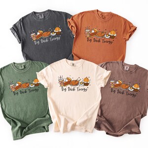May include: Five short-sleeved t-shirts in various colors: charcoal, orange, green, cream, and brown. Each shirt features a graphic of Thanksgiving food items and the text "Big Back Energy" in a script font. The shirts are made of a soft material.