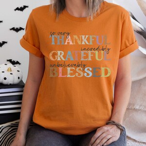 K&ouml;nnte beinhalten: Orangefarbenes T-Shirt mit dem Text "so very THANKFUL incredibly GRATEFUL unbelievably BLESSED" in bunten Buchstaben.