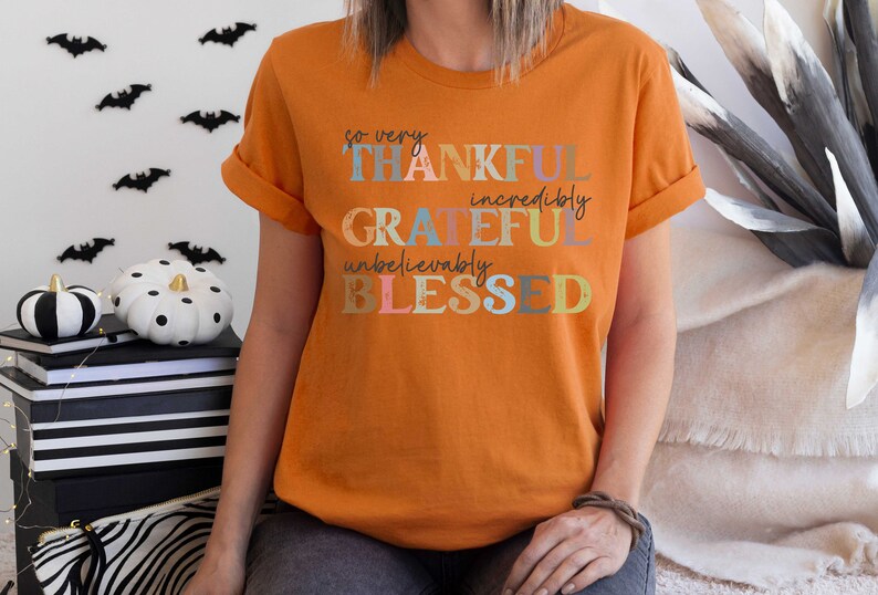 K&ouml;nnte beinhalten: Orangefarbenes T-Shirt mit dem Text "so very THANKFUL incredibly GRATEFUL unbelievably BLESSED" in bunten Buchstaben.