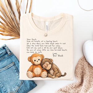 K&ouml;nnte beinhalten: Cremefarbenes T-Shirt mit einer Aquarellillustration von zwei Pl&uuml;schaffen. Einer sitzt, der andere schl&auml;ft. Das Shirt hat handgeschriebenen Text, der lautet: "Dear Punch, I am not breath..." und endet mit "Love, Your Plush."