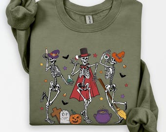 Può includere: Felpa verde oliva con un design a tema Halloween. La grafica presenta tre scheletri danzanti, ognuno con accessori festivi come cappelli, mantelli e una scopa. Pipistrelli, stelle, una lapide e una zucca completano il design.