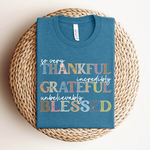 K&ouml;nnte beinhalten: Ein blaues T-Shirt mit dem Text "so very THANKFUL incredibly GRATEFUL unbelievably BLESSED" in bunten, abgenutzten Buchstaben.