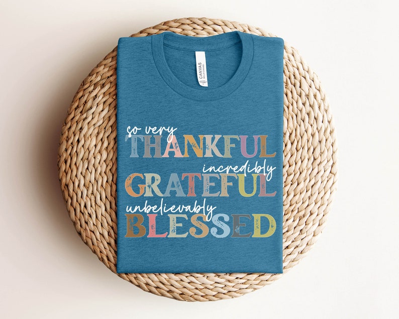 K&ouml;nnte beinhalten: Ein blaues T-Shirt mit dem Text "so very THANKFUL incredibly GRATEFUL unbelievably BLESSED" in bunten, abgenutzten Buchstaben.