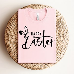 Puede incluir: Una camiseta rosa con el texto "Happy Easter" en letra negra con un gr&aacute;fico de oreja y cola de conejo.