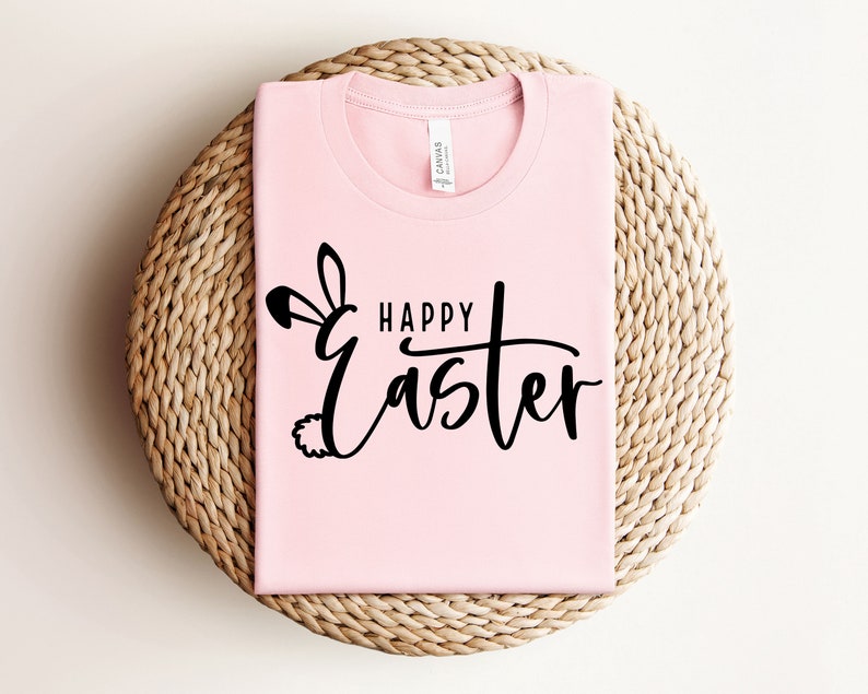 Puede incluir: Una camiseta rosa con el texto "Happy Easter" en letra negra con un gr&aacute;fico de oreja y cola de conejo.