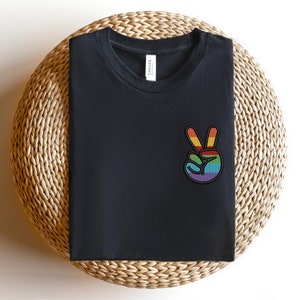 K&ouml;nnte beinhalten: Schwarzes T-Shirt mit einem Regenbogen-Friedenszeichen-Motiv. Das T-Shirt ist gefaltet und auf einer geflochtenen Strohmatte ausgestellt.