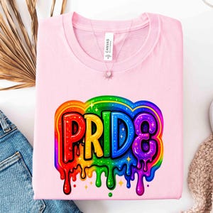 May include: A light pink t-shirt with the word "PRIDE" in a colorful, dripping rainbow design. The text is outlined in a darker color, with stars and drips adding to the design. The shirt is made of soft material.