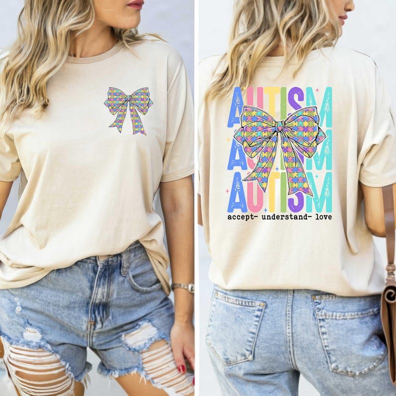 May include: Beige t-shirt with a colorful bow design on the front and back. The back of the shirt features the word "AUTISM" in large letters, along with the words "accept- understand- love". The bow design is made up of puzzle pieces.