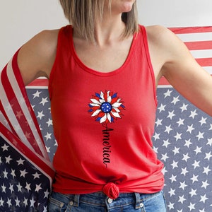Puede incluir: Un top rojo sin mangas con un dise&ntilde;o de girasol blanco, azul y rojo y la palabra "America" escrita verticalmente debajo. El top est&aacute; siendo sostenido por una persona que lleva pantalones vaqueros azules y una bandera estadounidense roja, blanca y azul drapeada sobre su hombro.