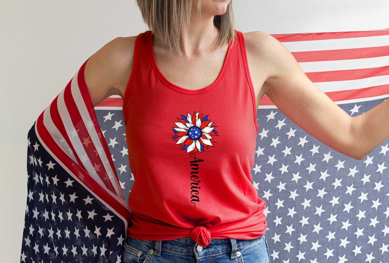Puede incluir: Un top rojo sin mangas con un dise&ntilde;o de girasol blanco, azul y rojo y la palabra "America" escrita verticalmente debajo. El top est&aacute; siendo sostenido por una persona que lleva pantalones vaqueros azules y una bandera estadounidense roja, blanca y azul drapeada sobre su hombro.