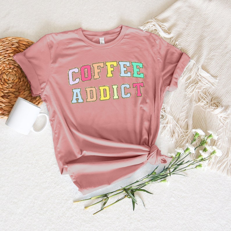 May include: A pink t-shirt with the text "COFFEE ADDICT" in colorful letters. The letters are outlined in gold glitter.