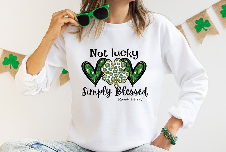 Puede incluir: Sudadera blanca con un tr&eacute;bol verde y dos corazones con el texto "Not lucky Simply Blessed Romans 4:7-8"