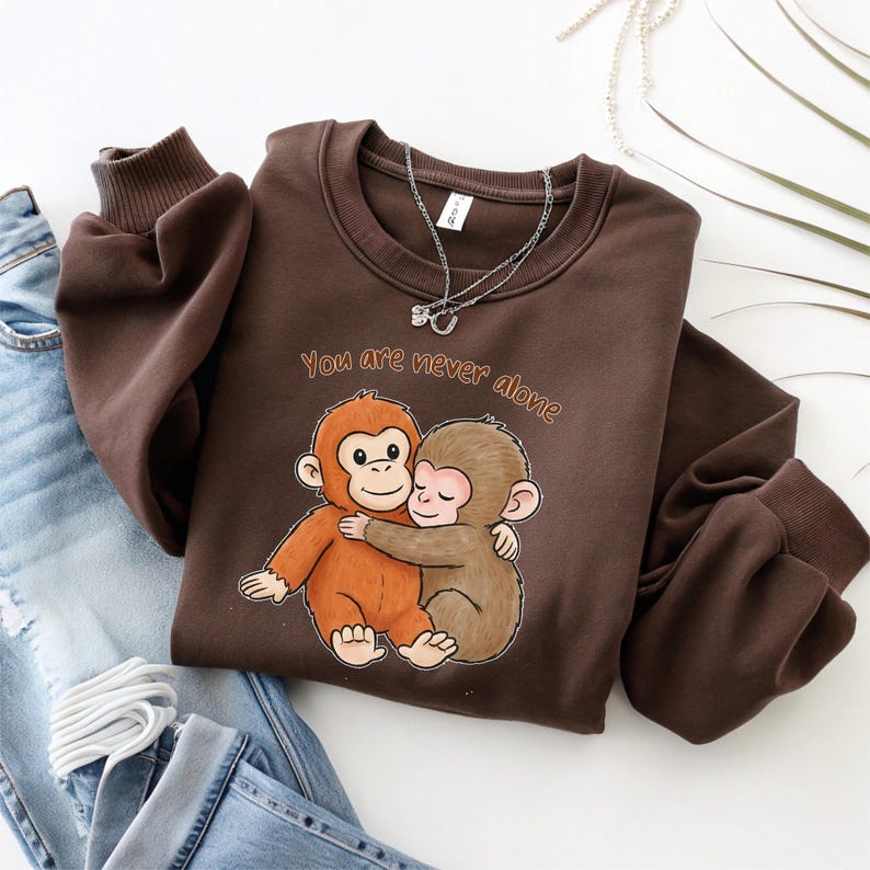 Puede incluir: Una sudadera marr&oacute;n con cuello redondo y mangas largas, con una ilustraci&oacute;n de dos monos abraz&aacute;ndose y el texto "You are never alone". Una cadena plateada est&aacute; drapeada sobre la parte superior de la sudadera.