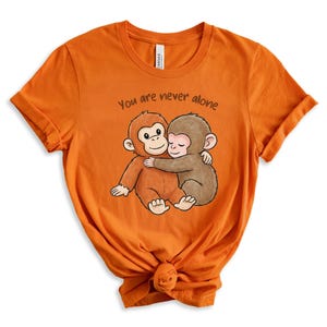 Puede incluir: Camiseta naranja con un gr&aacute;fico de dos monos de dibujos animados abraz&aacute;ndose. El texto "You are never alone" est&aacute; encima de los monos. La camiseta est&aacute; hecha de un material suave.