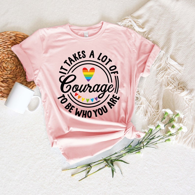May include: A light pink t-shirt with the text "It takes a lot of courage to be who you are" in a black circular design. The design includes a rainbow heart and rainbow hearts. The shirt is knotted at the bottom.