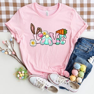 May include: Pink t-shirt with a graphic of a bunny rabbit and the word "LOVE" in colorful letters. The letters are decorated with Easter eggs, flowers, and a carrot.