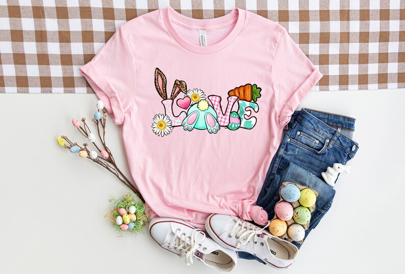 May include: Pink t-shirt with a graphic of a bunny rabbit and the word "LOVE" in colorful letters. The letters are decorated with Easter eggs, flowers, and a carrot.