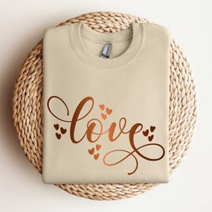 May include: A beige sweatshirt with the word "love" written in a cursive font with copper-colored hearts surrounding the word.