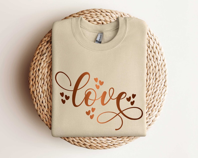 May include: A beige sweatshirt with the word "love" written in a cursive font with copper-colored hearts surrounding the word.
