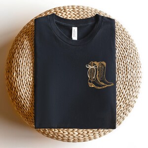 May include: Black t-shirt with a gold-colored illustration of cowboy boots. The boots are detailed with stitching and a bow. The shirt is displayed on a round, woven surface.