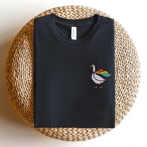 May include: Black t-shirt with a white goose wearing a rainbow cape. The goose is embroidered on the shirt.