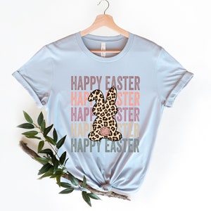 May include: Light blue t-shirt with the text "Happy Easter" repeated in a stacked design with a leopard print bunny in the center. The text is in a vintage style font with pink, orange, and yellow colors.