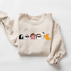 Pode incluir: Uma camisa de moletom bege com um gr&aacute;fico de cinco fantasmas, cada um representando um personagem de filme de terror diferente. Os fantasmas est&atilde;o em fila, e o &uacute;ltimo est&aacute; perseguindo um Pac-Man com uma faca.