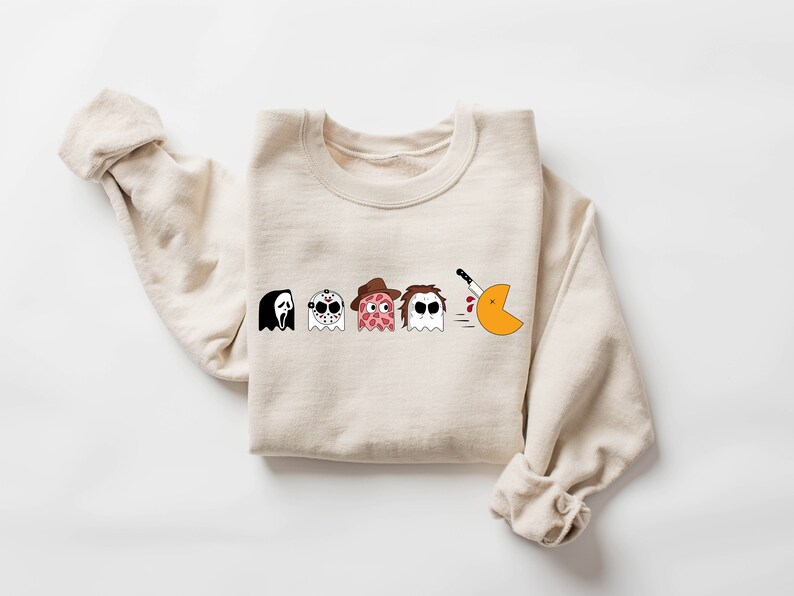Pode incluir: Uma camisa de moletom bege com um gr&aacute;fico de cinco fantasmas, cada um representando um personagem de filme de terror diferente. Os fantasmas est&atilde;o em fila, e o &uacute;ltimo est&aacute; perseguindo um Pac-Man com uma faca.