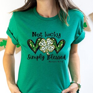 Puede incluir: Camiseta verde con un tr&eacute;bol de cuatro hojas y dos corazones, y el texto "Not lucky simply blessed Romans 5:7-8".