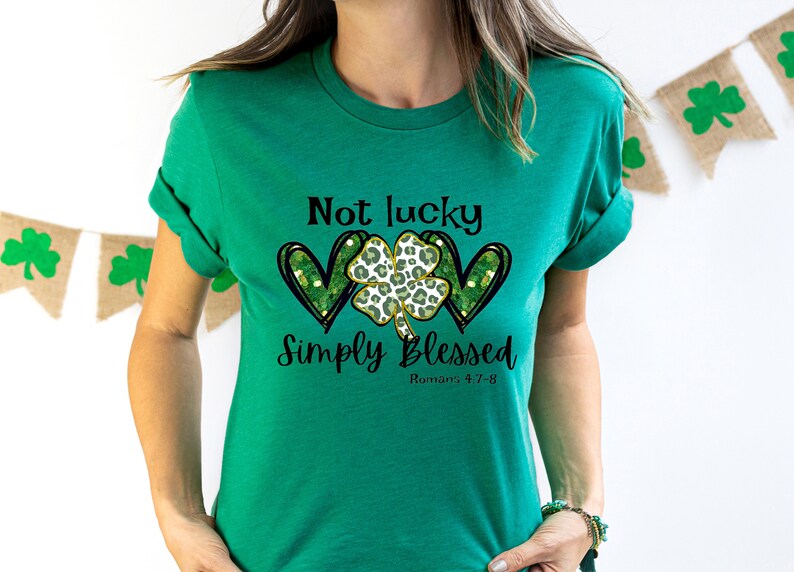 Puede incluir: Camiseta verde con un tr&eacute;bol de cuatro hojas y dos corazones, y el texto "Not lucky simply blessed Romans 5:7-8".