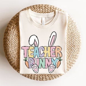 May include: A white t-shirt with a colorful graphic that says "Teacher Bunny" with bunny ears, paw prints, and carrots.