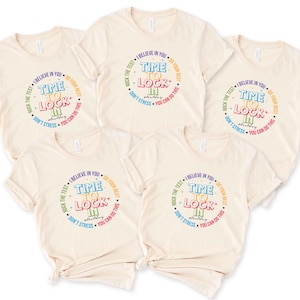 May include: Several cream-colored t-shirts with a circular graphic. The graphic reads "Time to Lock In" in colorful text, surrounded by phrases like "Believe in You" and "Do Your Best". The shirts are arranged on a white surface.