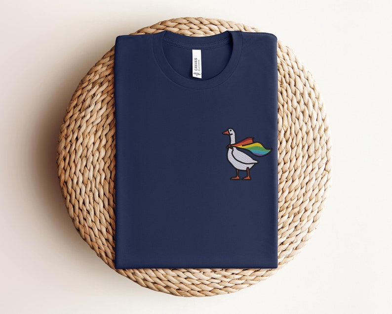 May include: A navy blue t-shirt with a white goose wearing a rainbow cape. The goose is facing right and has a black outline.
