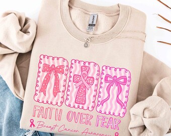 Könnte beinhalten: Beiges Sweatshirt mit rosa Grafiken für Brustkrebs-Aufklärung. Das Design umfasst drei rosa gerahmte Bilder einer Schleife, eines Kreuzes mit einem Band und einer Schleife. Der Text lautet "Faith Over Fear" und "Breast Cancer Awareness".