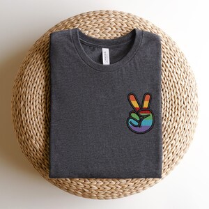 K&ouml;nnte beinhalten: Ein dunkelgraues T-Shirt mit einem gestickten Regenbogen-Friedenszeichen auf der linken Brustseite.
